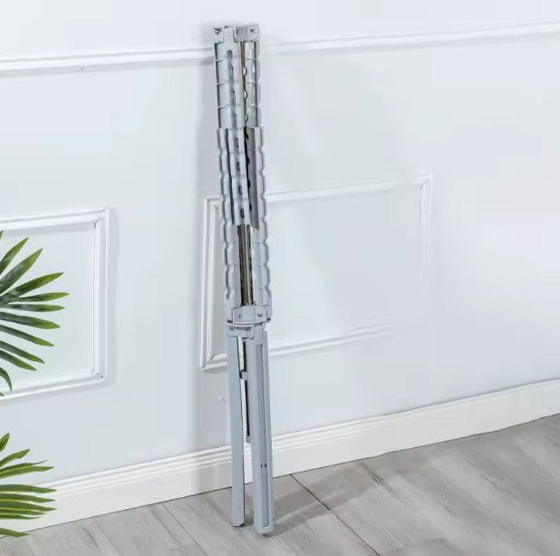 Tendedero de piso plegable metálico en tono gris claro, colapsado y apoyado verticalmente contra una pared blanca con molduras.