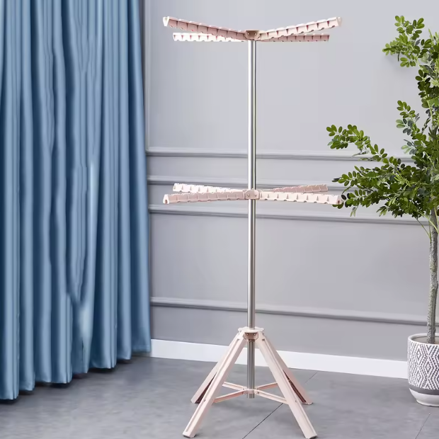 Tendedero de piso plegable con poste metálico y dos niveles de brazos rosados, ubicado en una habitación con cortina azul y planta decorativa.