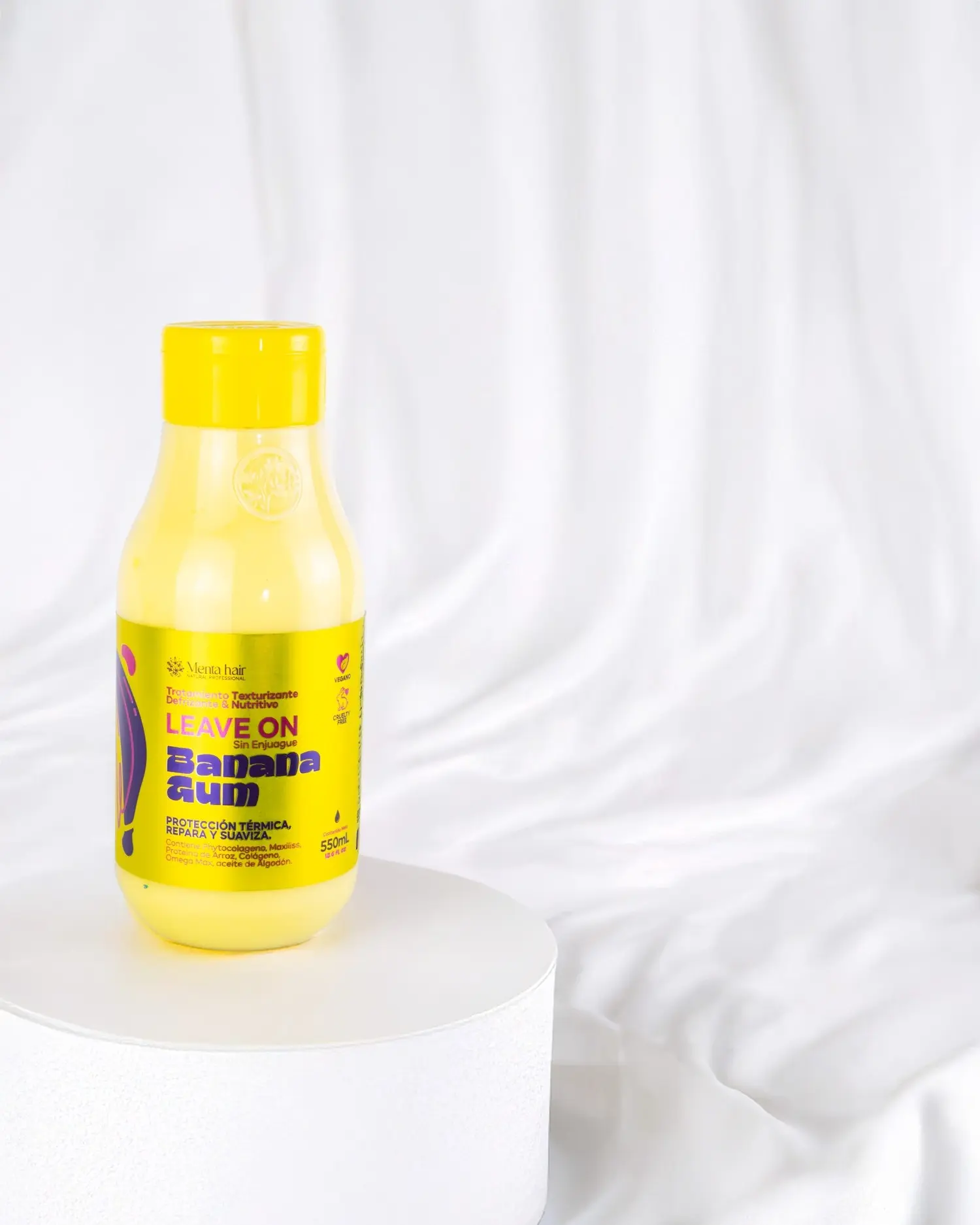 Crema para peinar termoprotectora Banana Gum en envase amarillo sobre pedestal blanco con fondo de tela blanca drapeada.