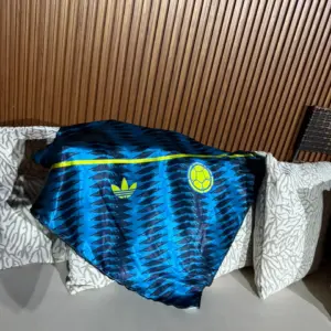 Pañoleta azul con patrón de plumas y detalles amarillos, con logo Adidas y escudo circular, sobre cojines blancos frente a pared de madera.