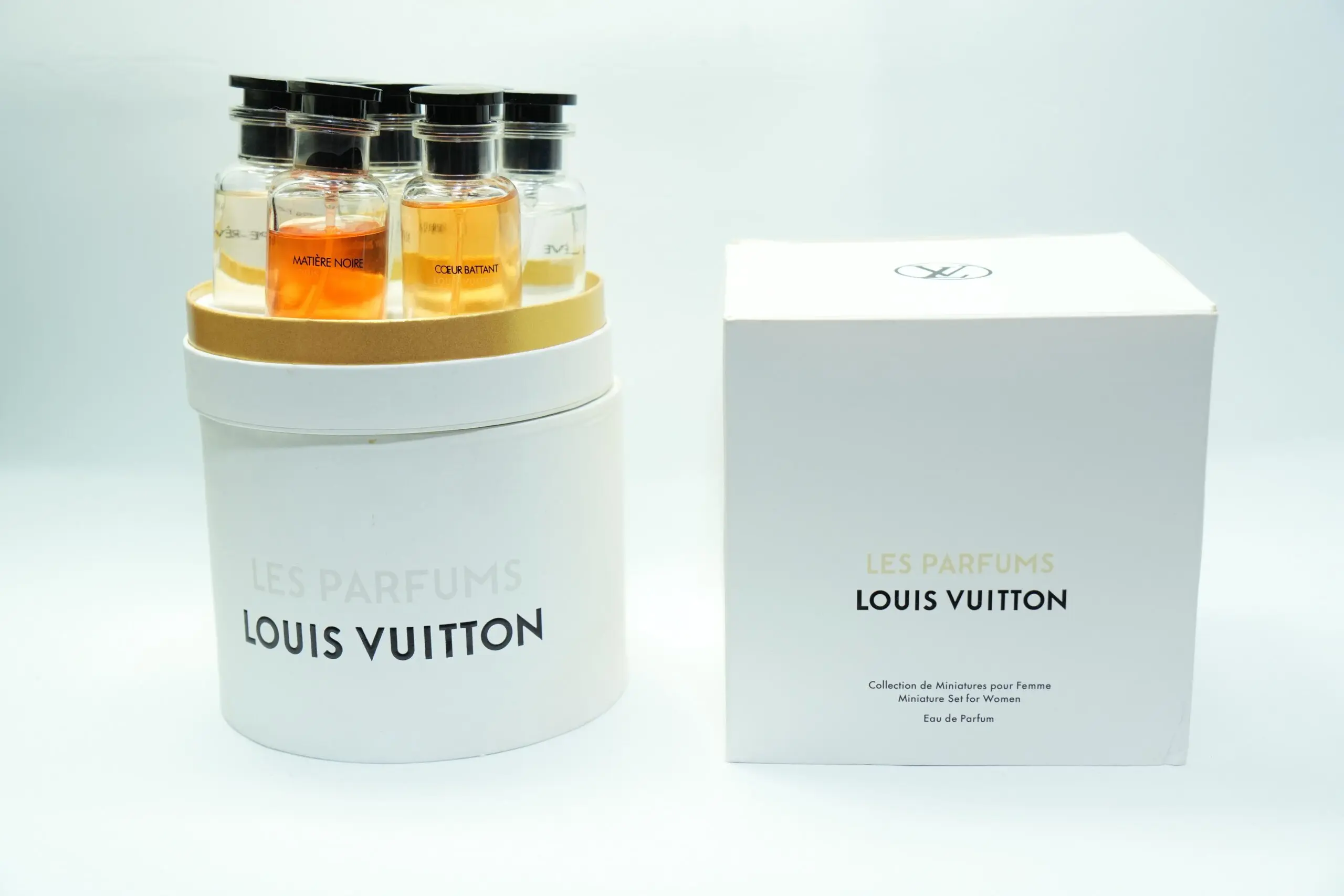 Set de miniaturas de perfumes Louis Vuitton en estuche cilíndrico blanco abierto con cinco botellas y caja de regalo al lado.