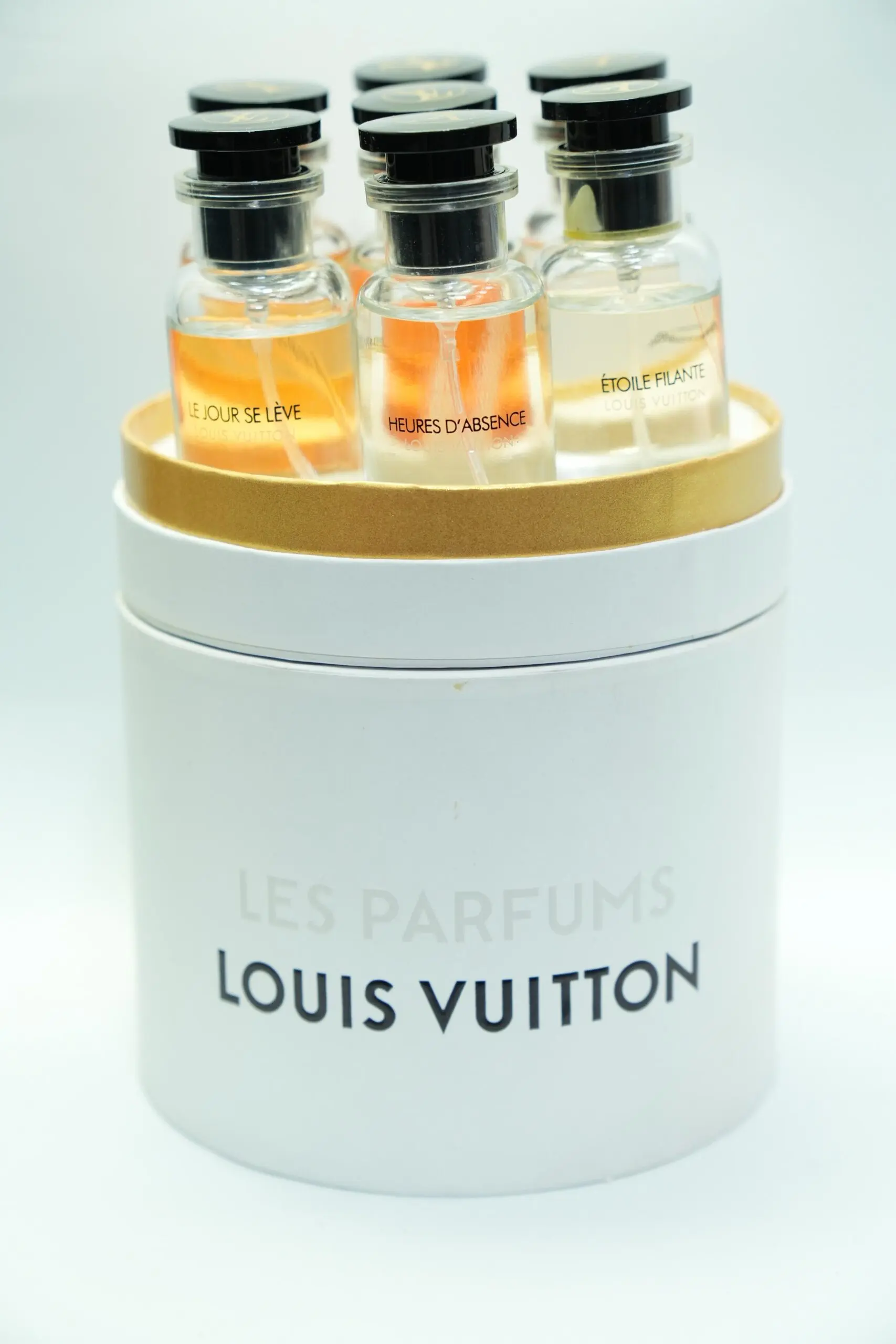 Estuche cilíndrico blanco y dorado de perfumes Louis Vuitton, que contiene varios frascos miniatura de vidrio con tapa negra y etiquetas visibles.
