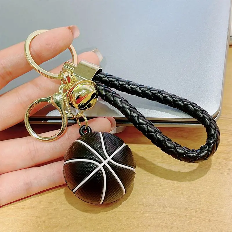 Llavero deportivo de baloncesto en miniatura negro con líneas blancas, correa trenzada de cuero sintético, argolla metálica dorada y cascabel.