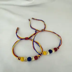 Set de dos manillas de hilo ajustable con cuentas de oro laminado y esferas de colores rojo, azul y amarillo sobre fondo blanco.