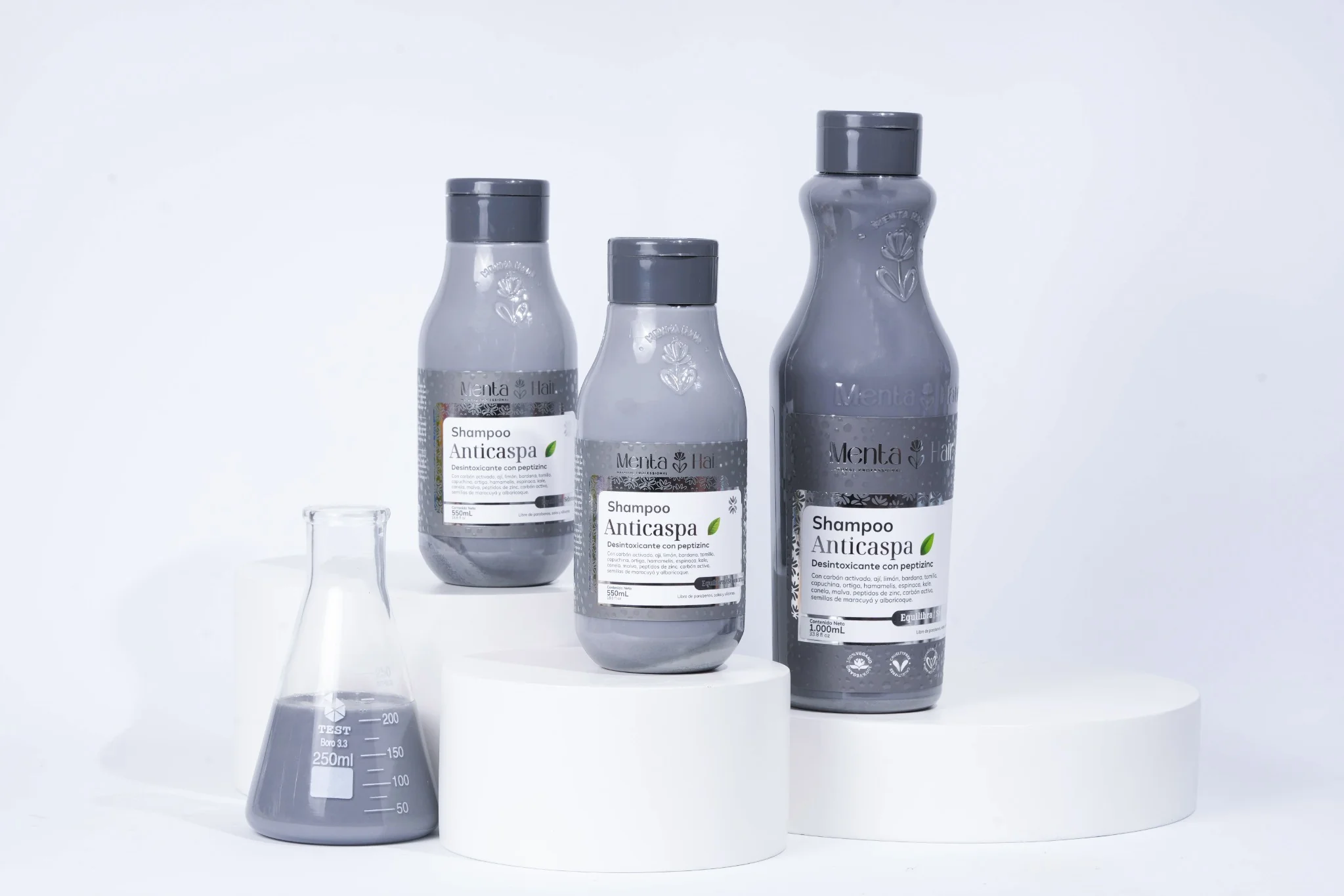 Tres botellas grises de shampoo anticaspa Menta Hair en tamaños variados, con matraz de laboratorio conteniendo líquido gris oscuro sobre pedestales blancos.