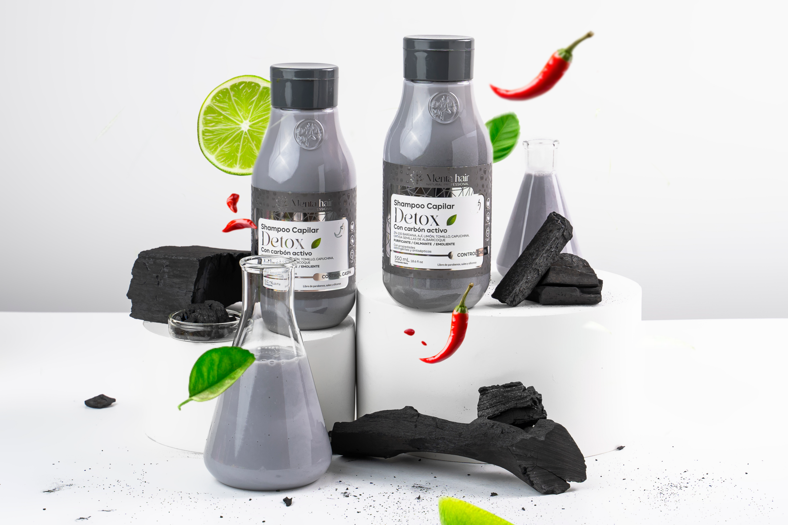 Shampoo capilar Detox con carbón activo en dos tamaños, acompañado de limones, chiles rojos, frascos de laboratorio y trozos de carbón vegetal.