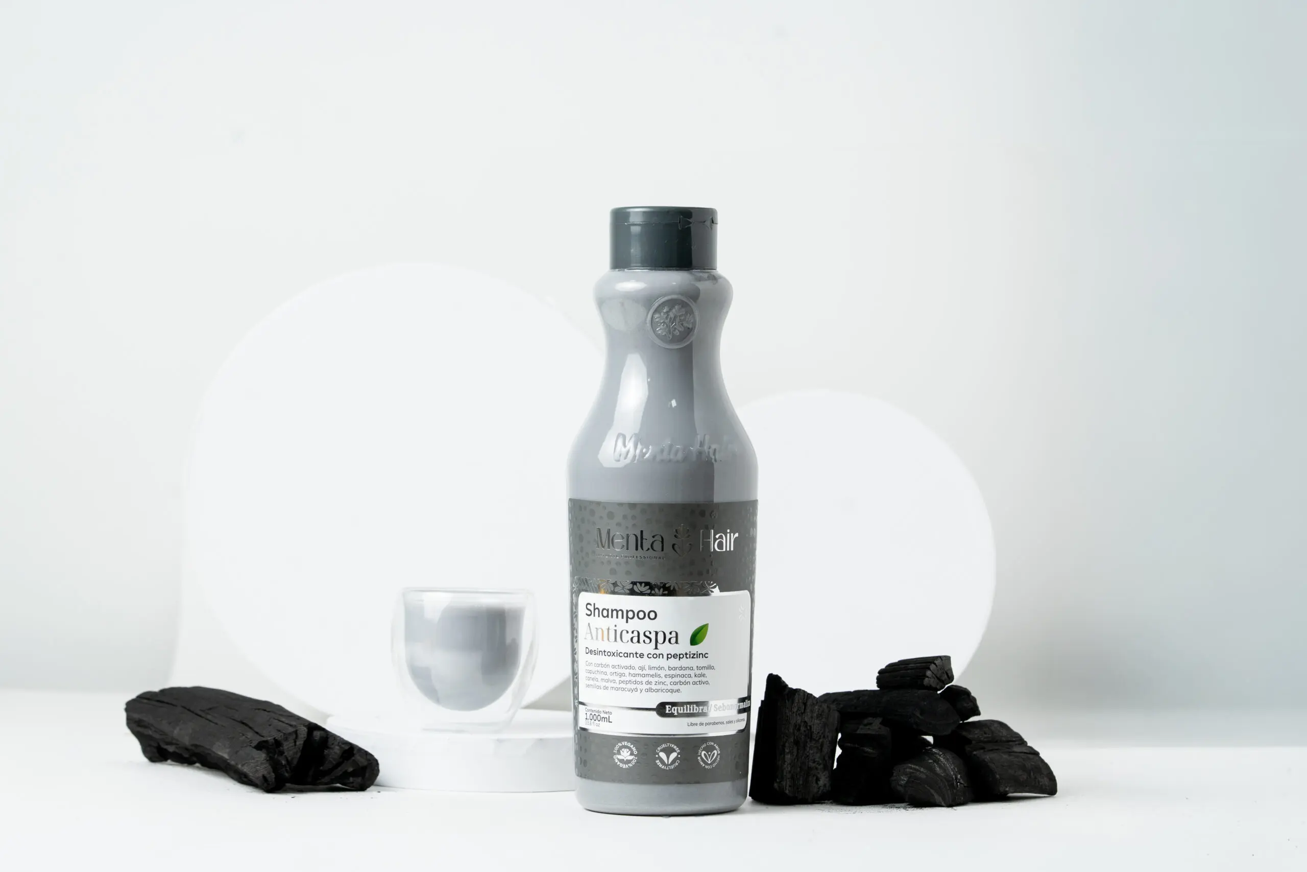 Shampoo anticaspa con carbón en botella gris de 1 litro, rodeado de trozos de carbón vegetal sobre un fondo blanco minimalista.
