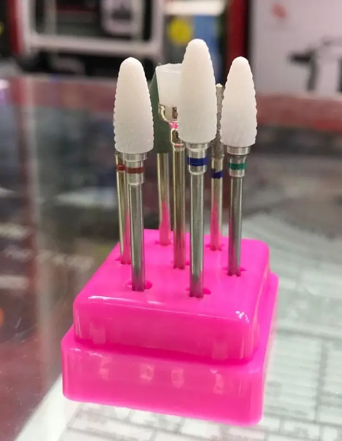 Set 7 brocas de cerámica para uñas en soporte rosa; incluye puntas cónicas y cilíndricas blancas, verdes y metálicas para manicura profesional.