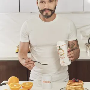 Hombre con bigote y moño sirve fibra de soya con psyllium e inulina en una taza junto a naranjas frescas y una torre de panqueques en la cocina.