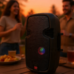Parlante portátil Bluetooth negro de 15 pulgadas con luz LED multicolor en la rejilla frontal, colocado sobre mesa de madera al aire libre con personas al fondo.