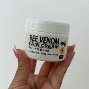 Mano sosteniendo un frasco blanco de 50 gramos de crema con veneno de abeja para articulaciones y huesos, con etiqueta de diseño hexagonal y abeja.