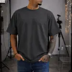 Hombre de perfil luciendo camiseta oversize gris oscuro de algodón con cuello redondo, corte relajado y jeans azules en estudio.