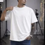 Hombre de piel morena con tatuajes viste camiseta oversize blanca de algodón con cuello redondo, en estudio de fotografía.