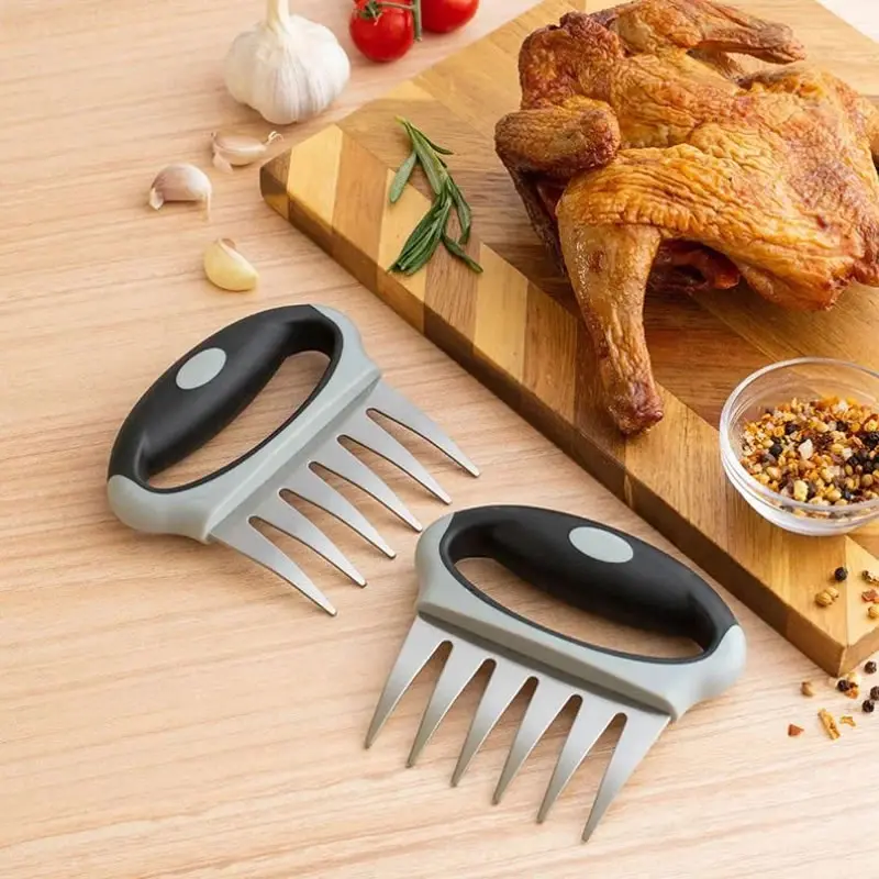 Par de garras trituradoras de carne estilo oso con mango ergonómico negro y gris, junto a pollo asado sobre tabla de madera.