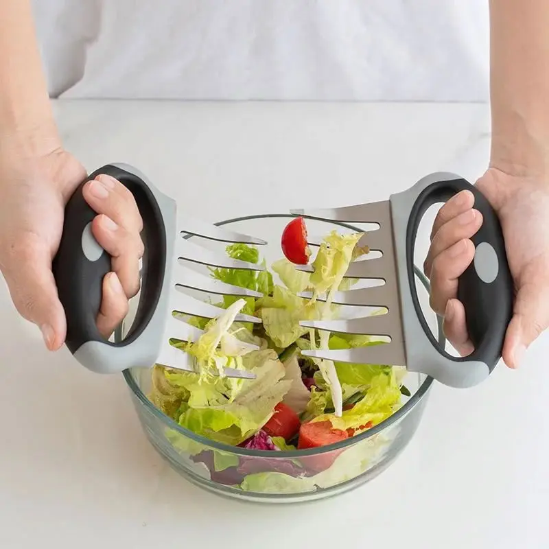 Persona usando dos garras trituradoras de carne grises y negras con mangos ergonómicos para mezclar ensalada en un bol de vidrio.