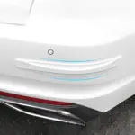Protector de parachoques blanco con detalles de líneas azules, instalado en la esquina de un vehículo blanco para evitar rayones y golpes.