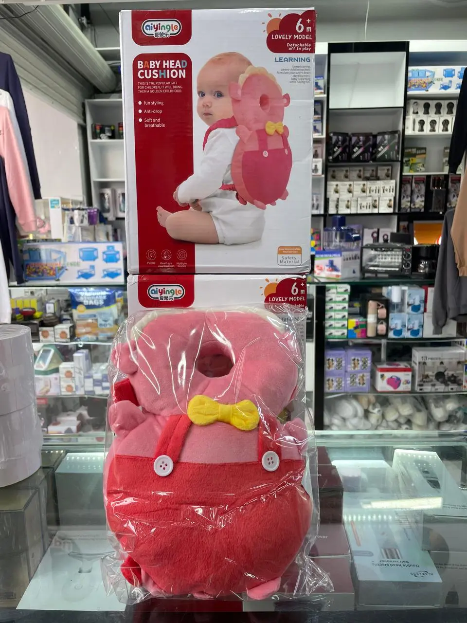 Cojín protector de cabeza rosa en forma de mochila para bebé, empaquetado debajo de su caja ilustrada sobre una vitrina de tienda.
