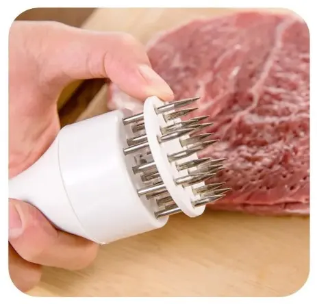 Ablandador de carne manual blanco con múltiples agujas de acero inoxidable, sostenido sobre una tabla de madera junto a un trozo de carne cruda.