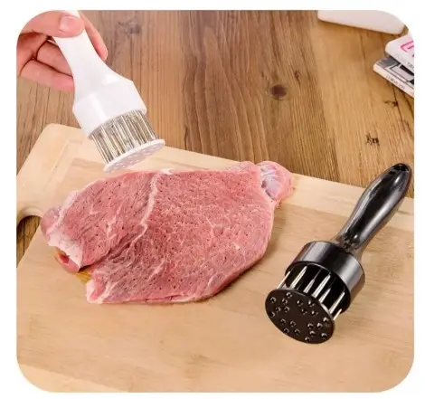 Set de dos ablandadores de carne manuales con mangos ergonómicos, uno blanco y otro negro, con cuchillas metálicas sobre tabla de madera y carne cruda.