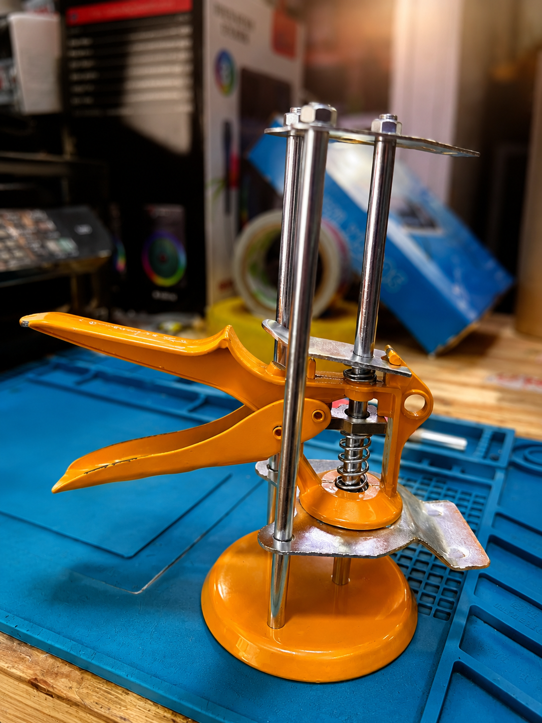 Herramienta de nivelación de enchapes color naranja con base circular, estructura metálica de dos guías y palanca de presión sobre tapete de trabajo azul.
