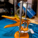 Herramienta de nivelación de enchapes naranja con base circular, ejes metálicos verticales y resorte, sobre tapete de trabajo azul para uso profesional.