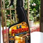 Botella negra con atomizador de limpieza La Patrona para pisos, con etiqueta de madera, sobre mesa blanca y fondo de jardín con plantas.