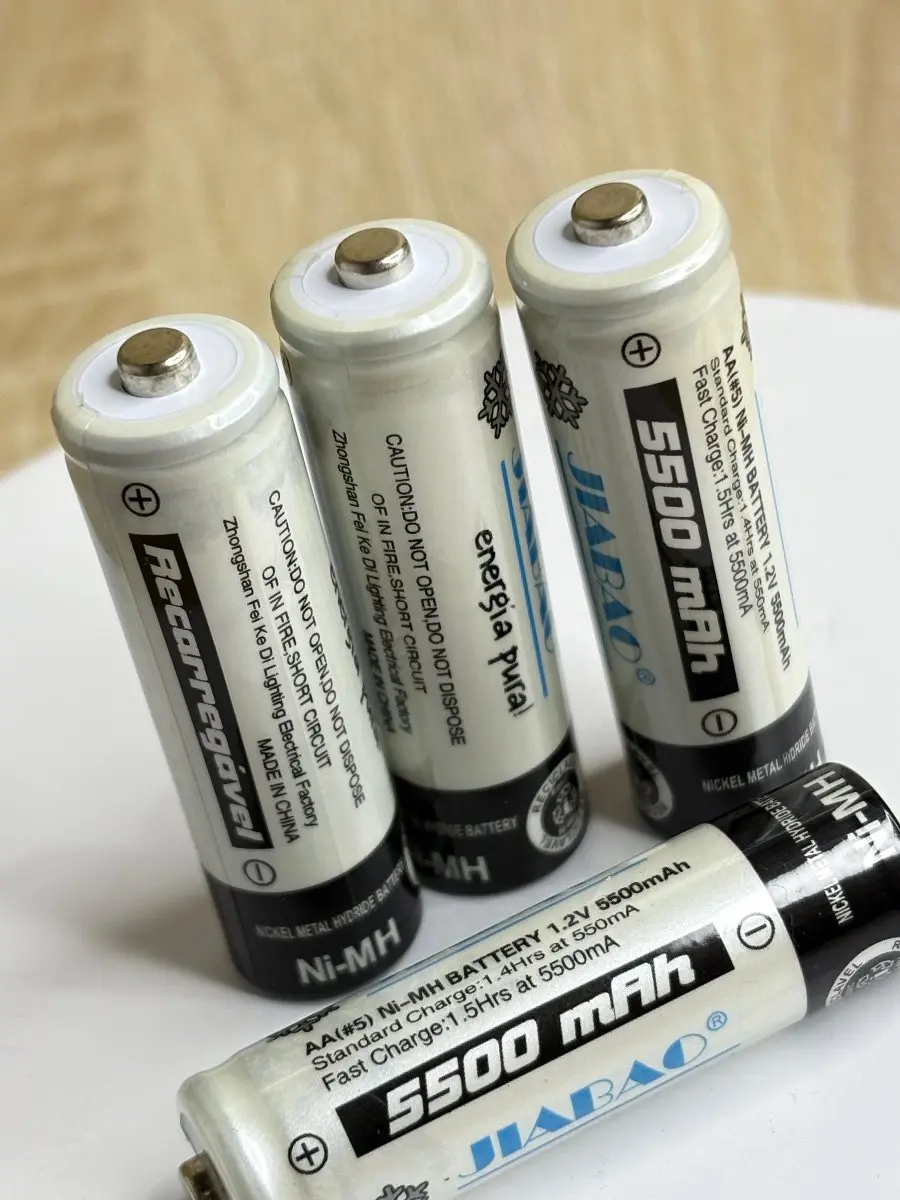 Cuatro pilas recargables AA Ni-MH blancas de 5500 mAh con detalles negros, una dispuesta horizontalmente y tres verticales sobre superficie blanca.