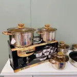 Batería de cocina de acero inoxidable brillante con bordes dorados, sobre caja negra, incluye cacerolas y ollas con tapas de vidrio y asas a juego.