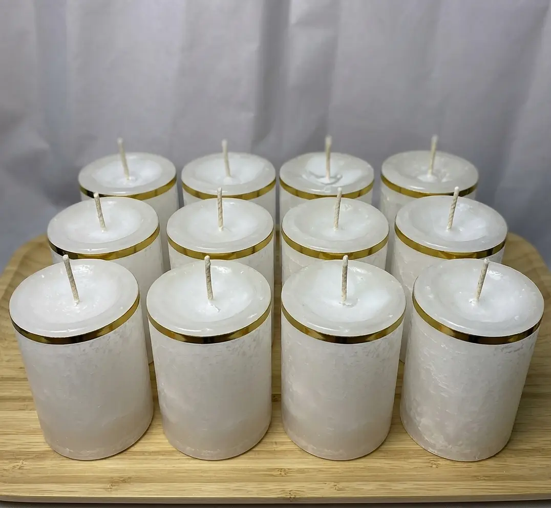 Doce veladoras artesanales de parafina blanca en envases cilíndricos con borde superior dorado, organizadas en cuatro filas sobre una bandeja de madera.