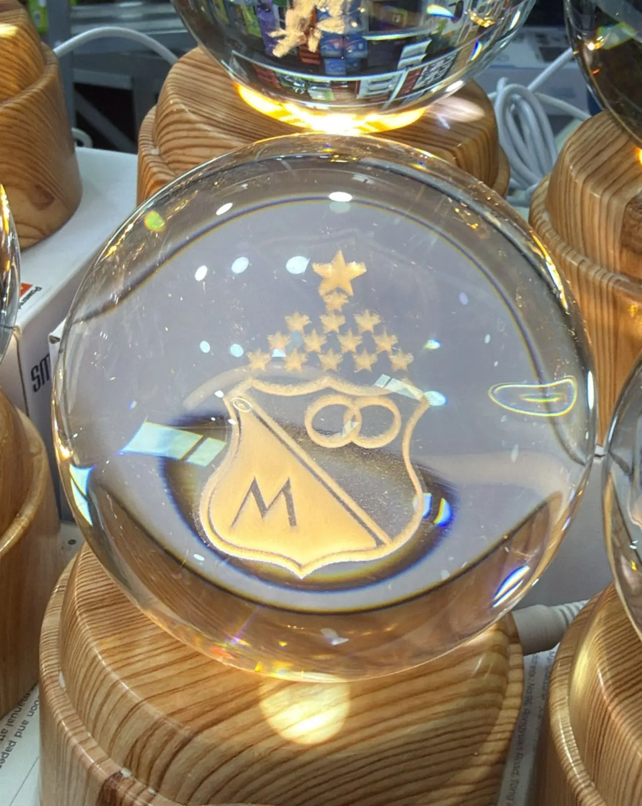Lámpara LED esférica de cristal con base de madera, grabada con el escudo del equipo Millonarios y estrellas, iluminada desde abajo sobre superficie.