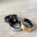 Anillos inteligentes de monitoreo biométrico en colores negro, plata y oro, con sensores internos visibles sobre una superficie de madera.