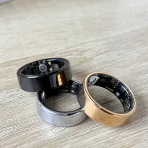 Anillos inteligentes de monitoreo biométrico en colores negro, plata y oro, con sensores internos visibles sobre una superficie de madera.