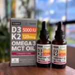 Set de dos frascos goteros de 30 ml de suplemento Nusava D3 K2 con Omega 3 y aceite MCT, sabor fresa, junto a su caja gris sobre mesa de madera.