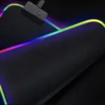 Tapete negro para escritorio con bordes de luces LED RGB en degradado multicolor, superficie lisa y conector USB visible en primer plano.
