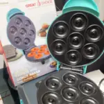 Máquina eléctrica para mini donas color turquesa con placas negras antiadherentes de siete moldes abiertas, junto a su caja de empaque.