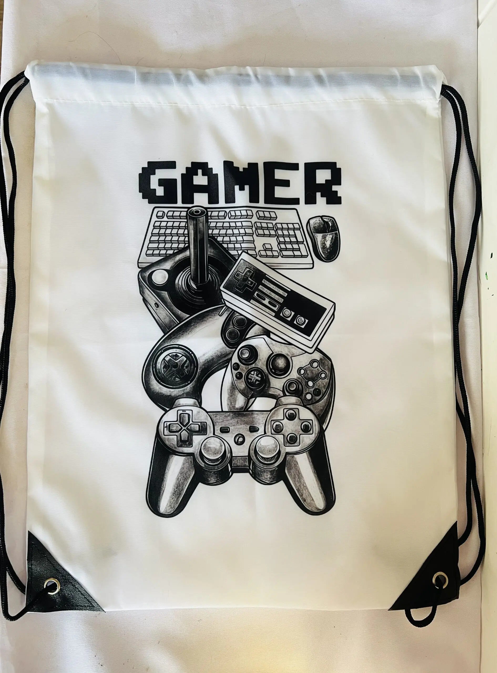Mochila saco blanca con cordones negros, diseño estampado de mandos retro, teclado y la palabra gamer.