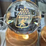 Lámpara LED esférica de cristal con escudo grabado del América de Cali, base circular de madera clara y cable de alimentación, ideal para decoración.