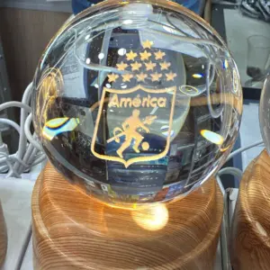 Lámpara LED esférica de cristal con escudo grabado del América de Cali, base circular de madera clara y cable de alimentación, ideal para decoración.
