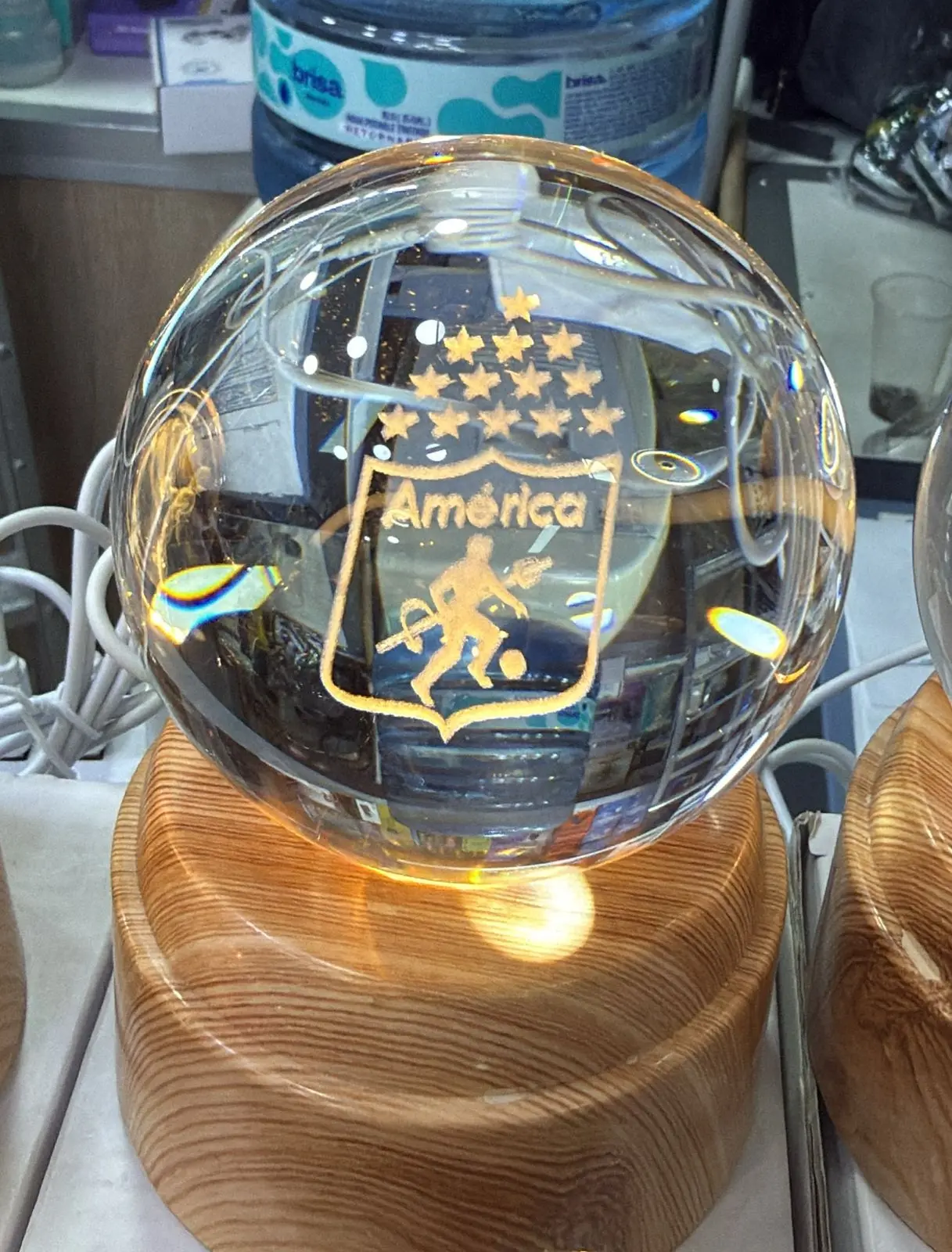 Lámpara LED esférica de cristal con escudo grabado del América de Cali, base circular de madera clara y cable de alimentación, ideal para decoración.