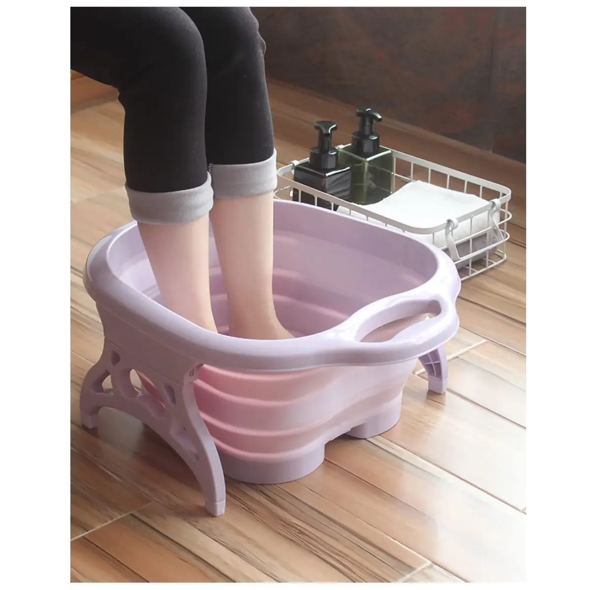 Tina spa plegable para pies en tonos lavanda y rosa con diseño flexible y asas, en uso sobre un piso de madera.