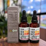 Set de dos frascos de suplemento líquido Nusava D3 K2 Omega 3 con aceite MCT, sabor fresa, junto a su caja con tabla nutricional sobre mesa de madera.