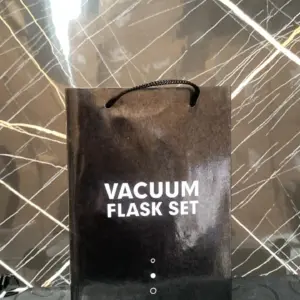 Bolsa de regalo de cartón negro brillante con el texto en blanco Vacuum Flask Set, asas negras y fondo texturizado.