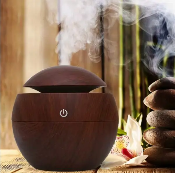 Difusor de aromaterapia ultrasónico oscuro con acabado efecto madera oscura, forma esférica y botón de encendido frontal, emitiendo vapor junto a piedras zen y una flor blanca.
