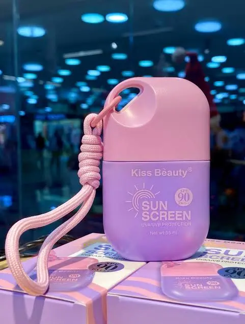 Protector solar facial en envase morado y tapa rosada con cordón de tela, fórmula FPS 90, sobre cajas de presentación.