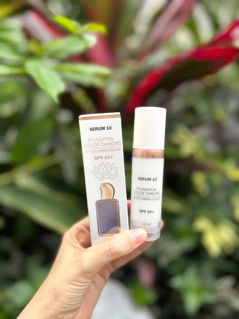 Mano sosteniendo caja y botella de base de maquillaje Serum 10 con SPF 50 frente a un fondo difuminado de hojas verdes y rojas.