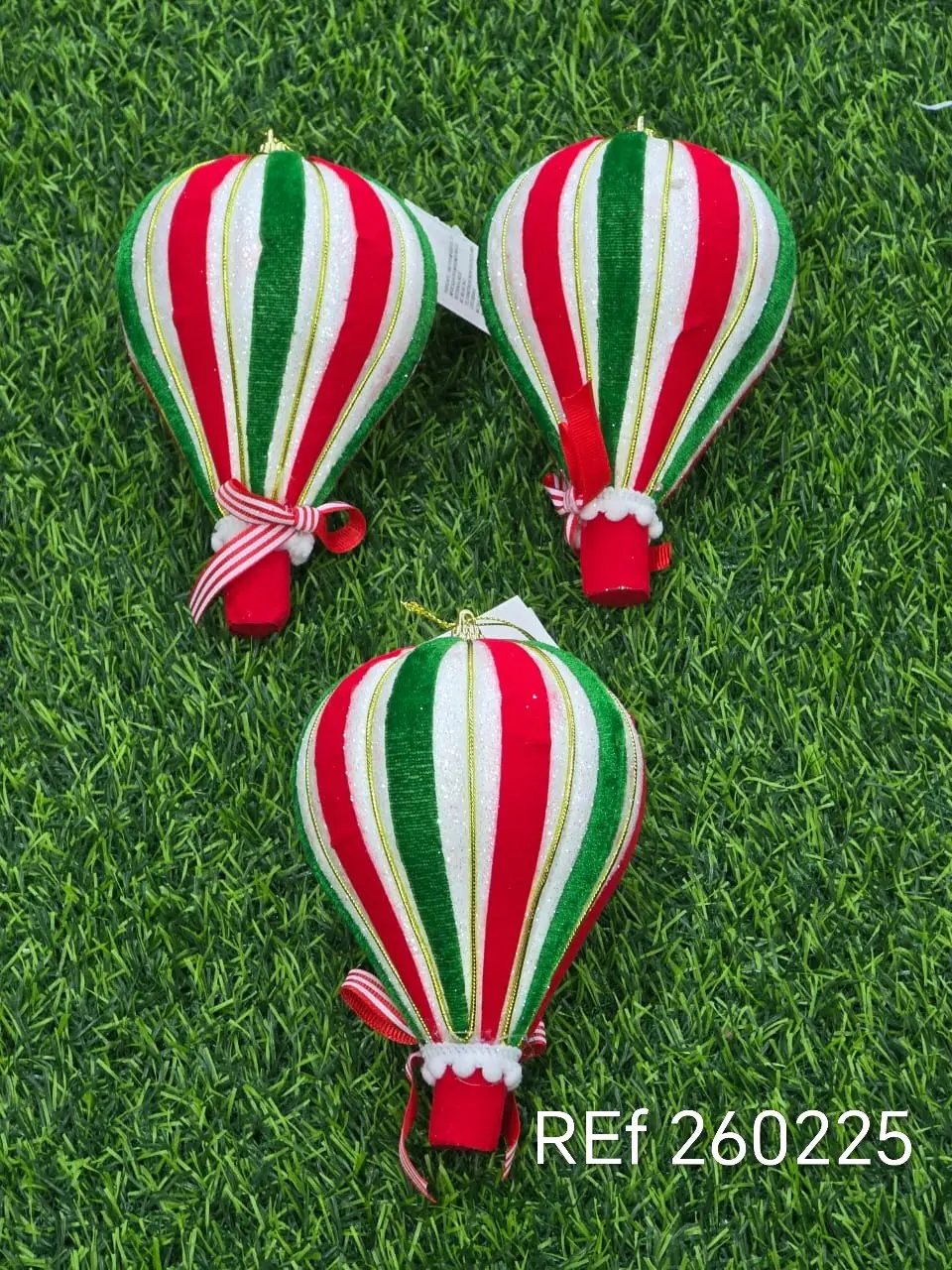 Adornos navideños colgantes de globos aerostáticos con rayas rojas, verdes y blancas y lazos decorativos sobre fondo de césped verde.