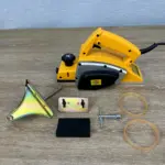 Cepilladora eléctrica amarilla para madera sobre superficie de madera, acompañada de guía paralela, dos correas de transmisión, soporte metálico y llave de ajuste.