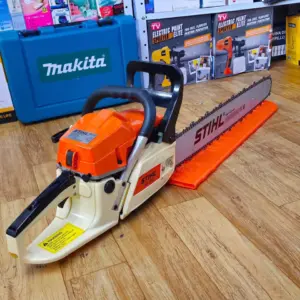 Motosierra a gasolina Stihl naranja y crema con espada larga y protector, apoyada sobre piso de madera en un almacén de herramientas.