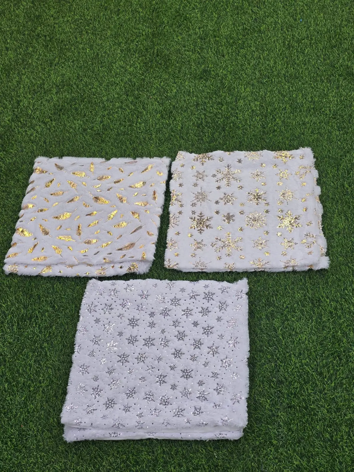 Tres cojines de pelo sintético blanco con estampados de hojas doradas, copos de nieve y estrellas plateadas sobre césped artificial verde.