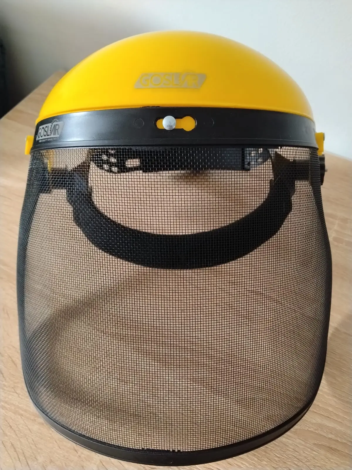Careta de protección facial para guadañadora con casco amarillo y visor de malla negra, vista frontal sobre superficie de madera.